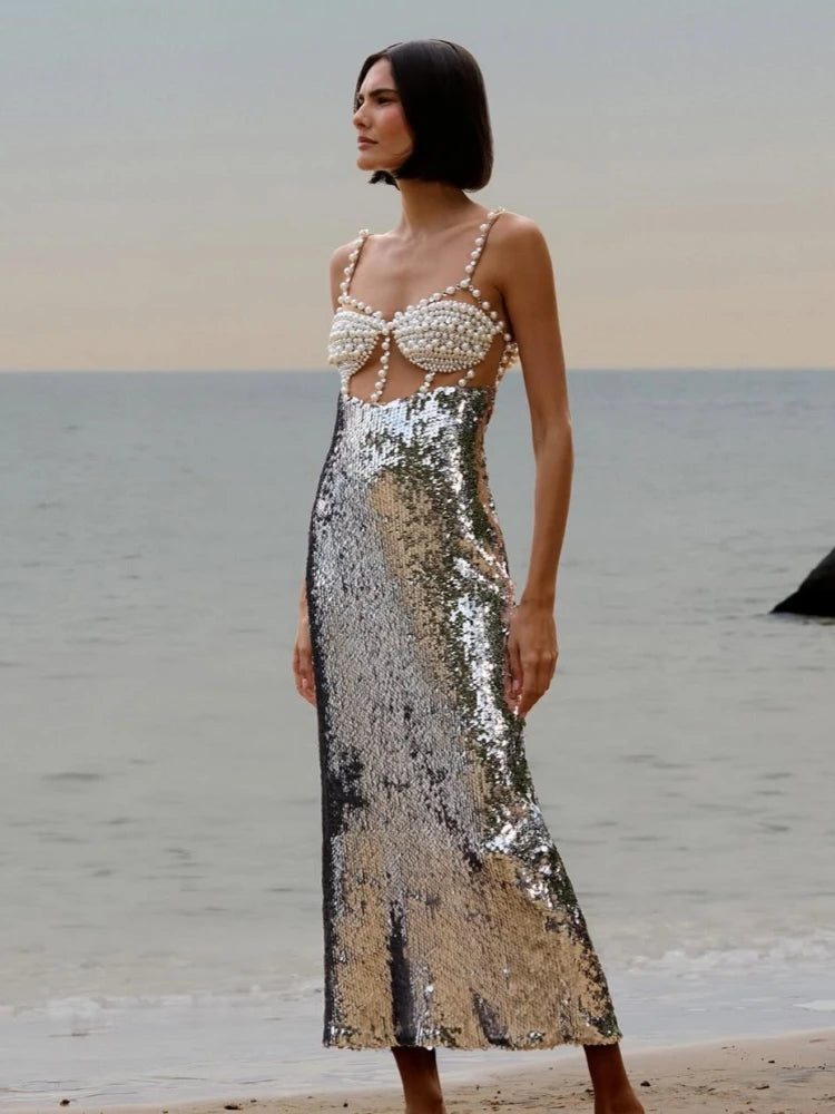 Model wearing a silver beaded sequin midi dress with spaghetti straps, standing along a shoreline at sunset.