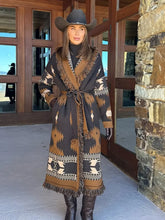 Full-length shot of a woman wearing a brown printed belted knit cardigan with tassel trim, styled with tall boots outside stone walls.