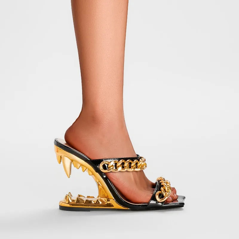On-foot studio shot of black and gold peep-toe mules with chunky chain strap, highlighting the sharp, sculptural heel profile.