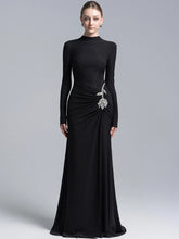 Model wearing a black high neck long sleeve maxi gown with draped waist and crystal floral detail, photographed on a clean studio background