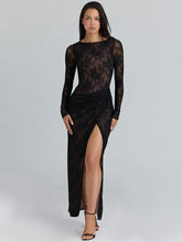Model in a black long sleeve lace maxi dress with a high slit, standing in heels against a clean studio gray background.