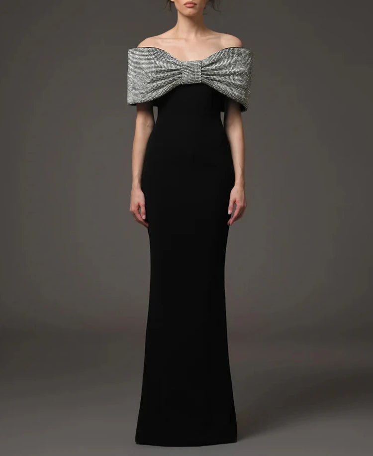 Front view of a black off-shoulder maxi dress featuring a sculptural rhinestone bow neckline against a dark studio backdrop.