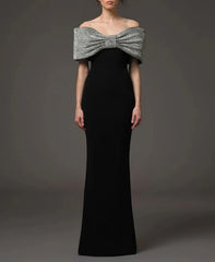 Front view of a black off-shoulder maxi dress featuring a sculptural rhinestone bow neckline against a dark studio backdrop.