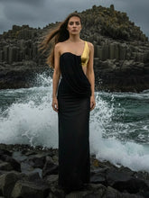 Model in a black one-shoulder maxi dress with a gold accent, standing on rocks as ocean waves crash behind