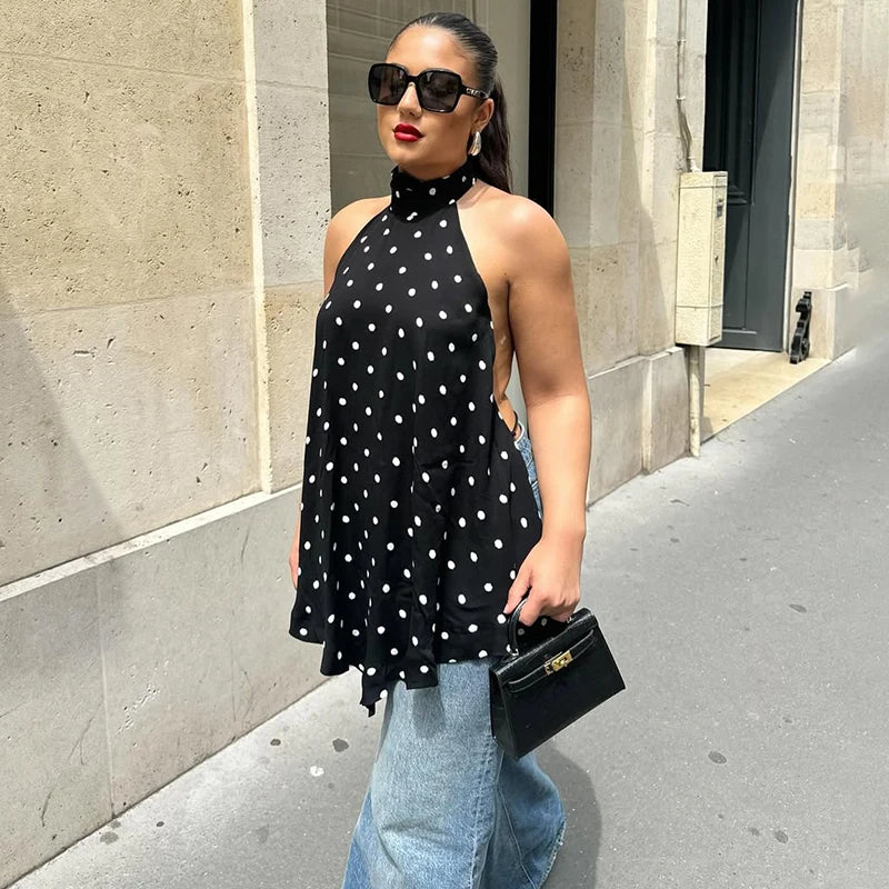 Woman wearing a black polka dot halter backless top styled for street summer fashion and blue jeans, standing on a sidewalk.