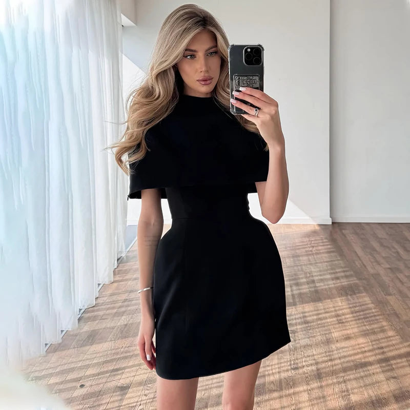 Front view of a black round-neck mini dress featuring a structured cape layer that frames the upper body with clean lines.