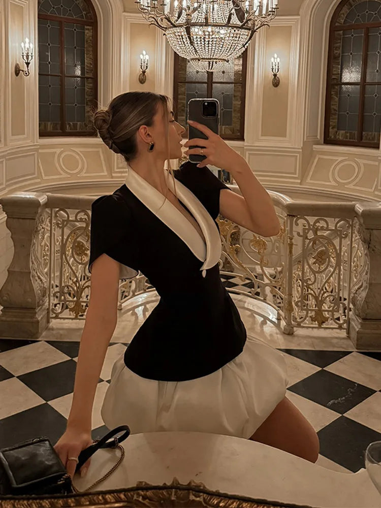 Mirror selfie wearing a black fitted top with a contrasting V-neck trim and a light mini skirt in an elegant chandelier-lit interior.