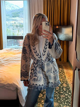 Mirror selfie in a room, wearing a blue-and-ivory paisley knit cardigan tied at the waist with fringe hem over denim.