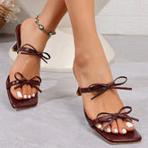 Brown bow square toe high heel mules worn with white nail polish for a summer outfit, worn by a person on a light background