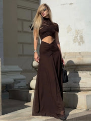 Full-length view shows the short sleeves, round neckline, and body-skimming ruched silhouette of the brown ankle-length dress in warm light.