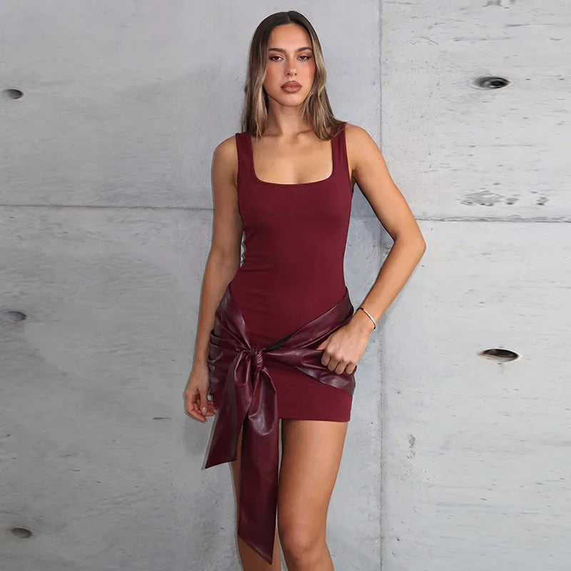 Woman wearing a burgundy square neck mini dress against a concrete wall, styled with a dark faux-leather ribbon tied at the hips.