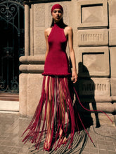 Full-length street shot of a burgundy halter maxi dress with long tassel fringe, styled against a stone building in bright sunlight.