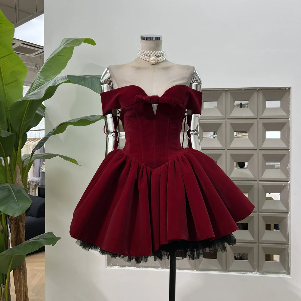 Mannequin in a burgundy velvet off-shoulder corset mini dress with a bow and pleated puff skirt in a studio setting.