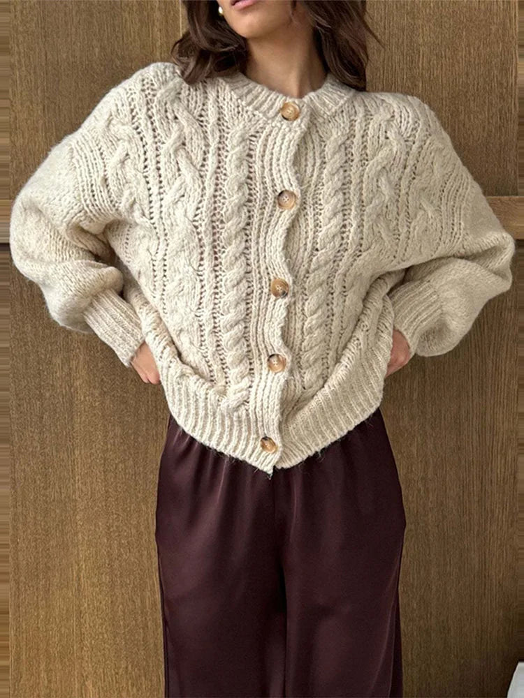 Beige cable-knit cardigan with button front styled with satin-finish trousers, showing the relaxed shape and textured knit in warm indoor light.