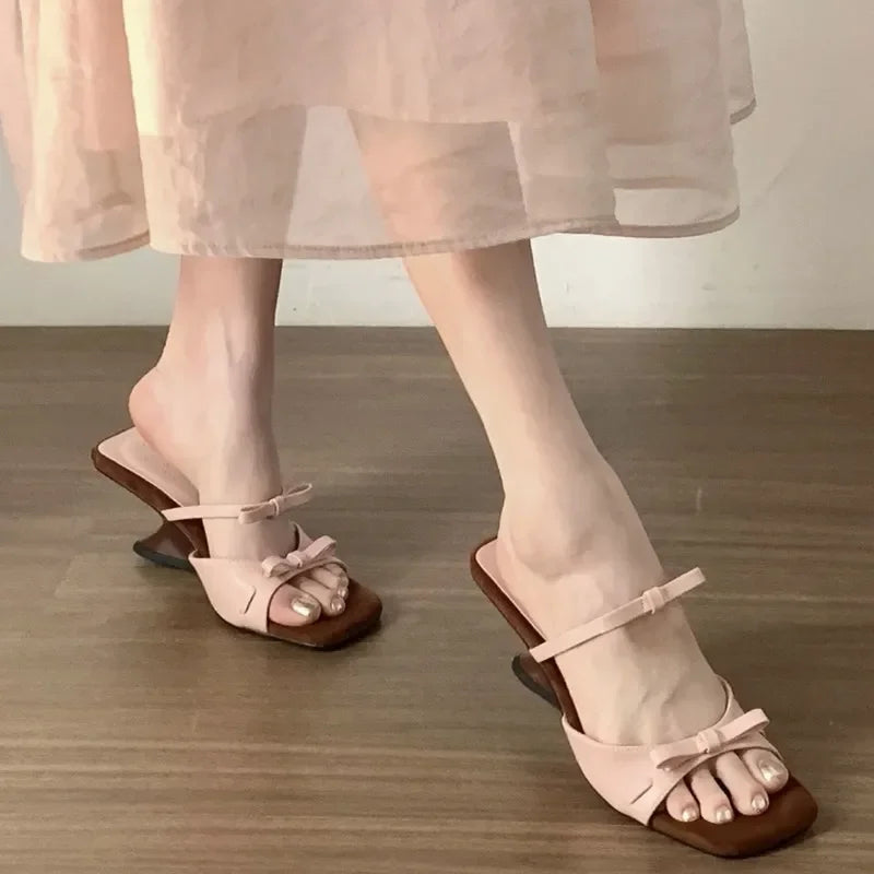 Pink sandals with brown soles worn by a person in a light dress on a wooden floor.
