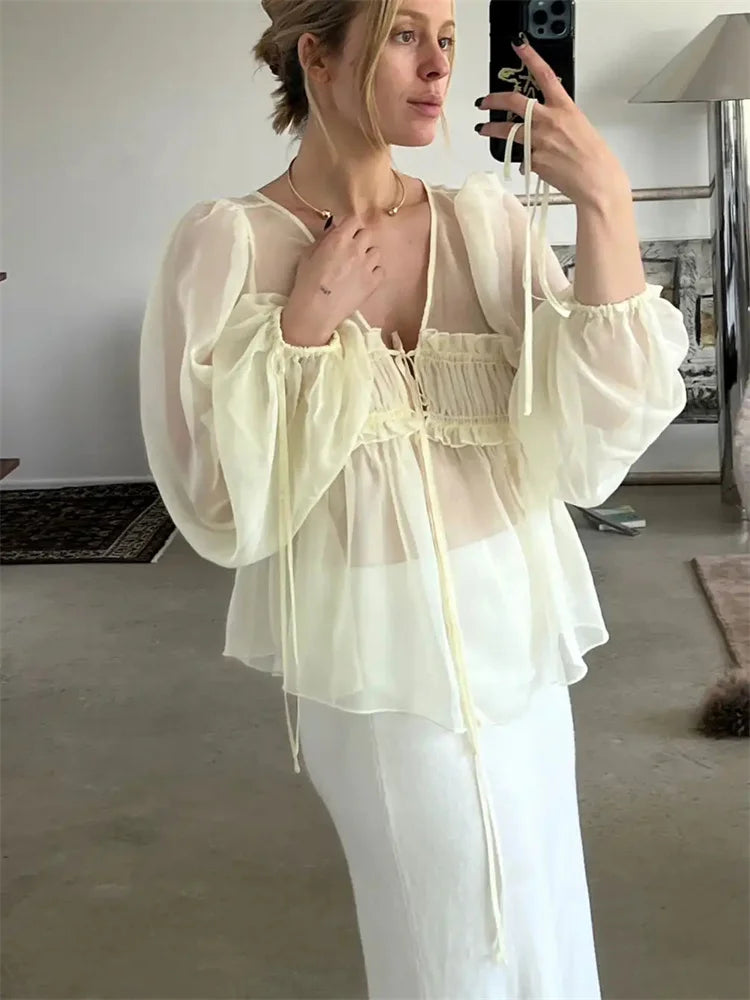 Woman taking a mirror selfie wearing a sheer top and white pants in a room.