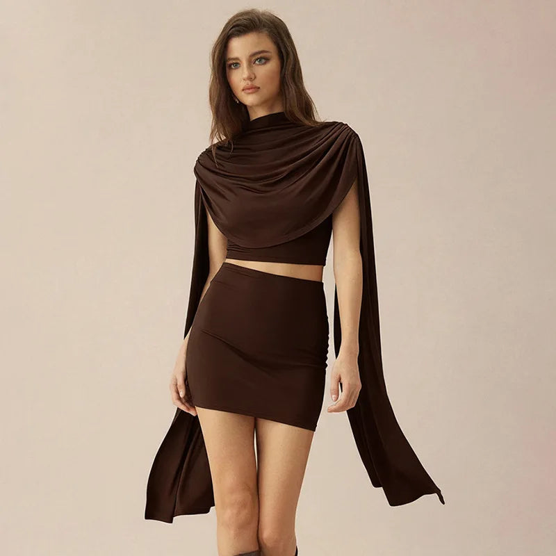 Model wearing a chocolate-brown draped shawl top with scarf detail and matching fitted mini skirt on a neutral background.