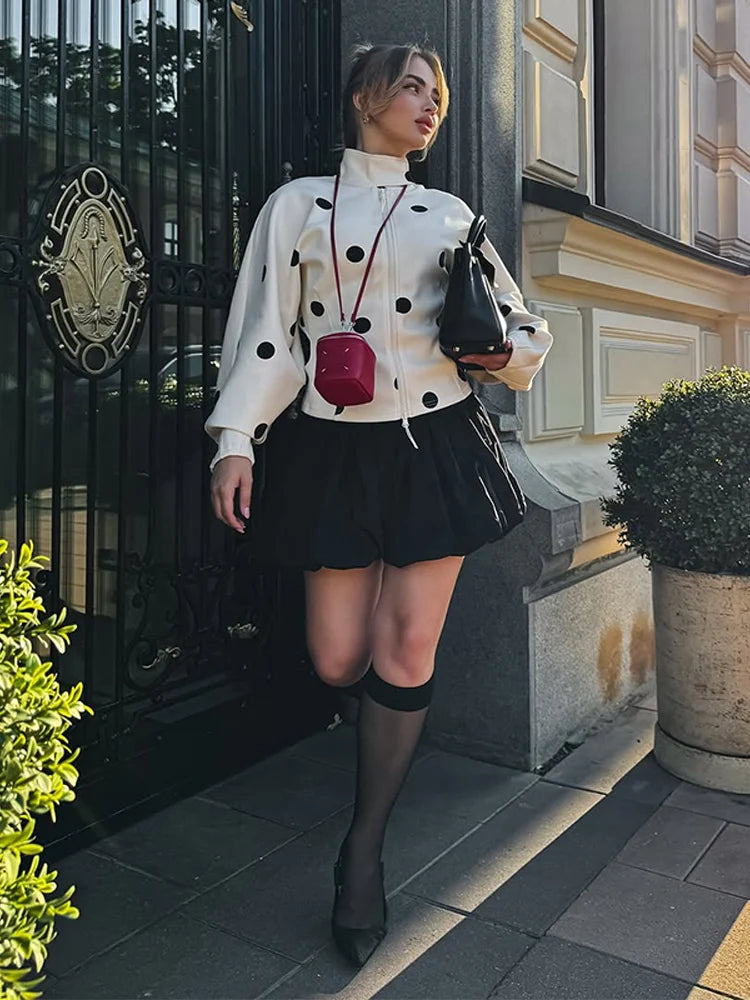A woman in a polka dot blouse and black skirt standing in front of a building entrance.