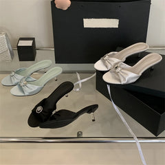 Crystal bow slides shown in a reflective setting for outdoor celebrations. Three pairs of high-heeled shoes in black, white, and light blue on a reflective surface with a black box in the background.