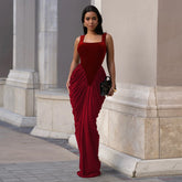 Model in a deep red velvet maxi dress with a square neckline standing by classical stone architecture holding a small clutch.