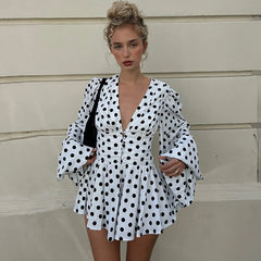 A woman wearing a deep V-neck high waist dot dress, shown with a black bag and boots against a beige wall.