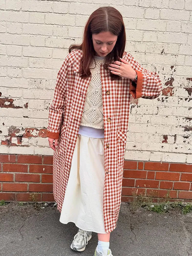 Person wearing a red and white checkered coat over a white dress, standing against a brick wall.