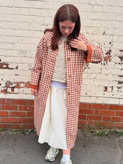 Person wearing a red and white checkered coat over a white dress, standing against a brick wall.