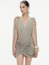 Model wearing a taupe draped mini dress with cape sleeves and a deep V-neck, styled with gold earrings on a clean studio backdrop.