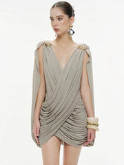Model wearing a taupe draped mini dress with cape sleeves and a deep V-neck, styled with gold earrings on a clean studio backdrop.