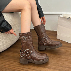 Women’s block heel mid-calf boots with decorative metal buckles worn by a person sitting on a wooden floor.