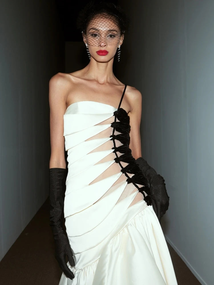 Editorial luxury gala maxi gown in white with dramatic glove accent. Model wearing a white dress with black lace details and black gloves indoors.