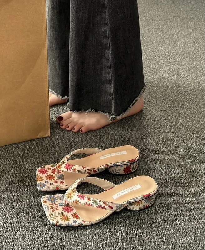 Elegant beach slippers with a flower print are shown for casual parties on a grey carpet with a cardboard box in the background.