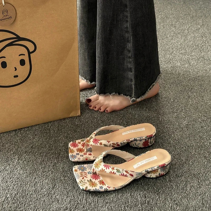 Elegant beach slippers with a flower print are shown for casual parties on a grey carpet with a cardboard box in the background.