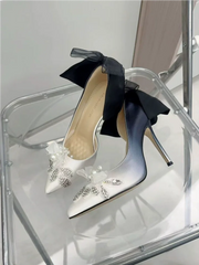 White high-heeled shoes with black bows and embellishments on a clear stand.