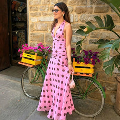 Woman in a pink polka dot dress standing next to a green bicycle with flower crates, against a stone wall.