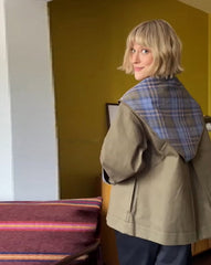 Person wearing a plaid shirt and beige coat in a room with a yellow wall and ironing board.
