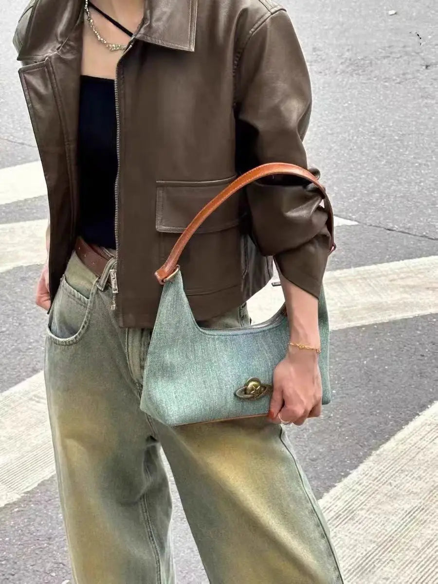 Person holding a light gold emblem detail on a retro denim underarm handbag on a street.