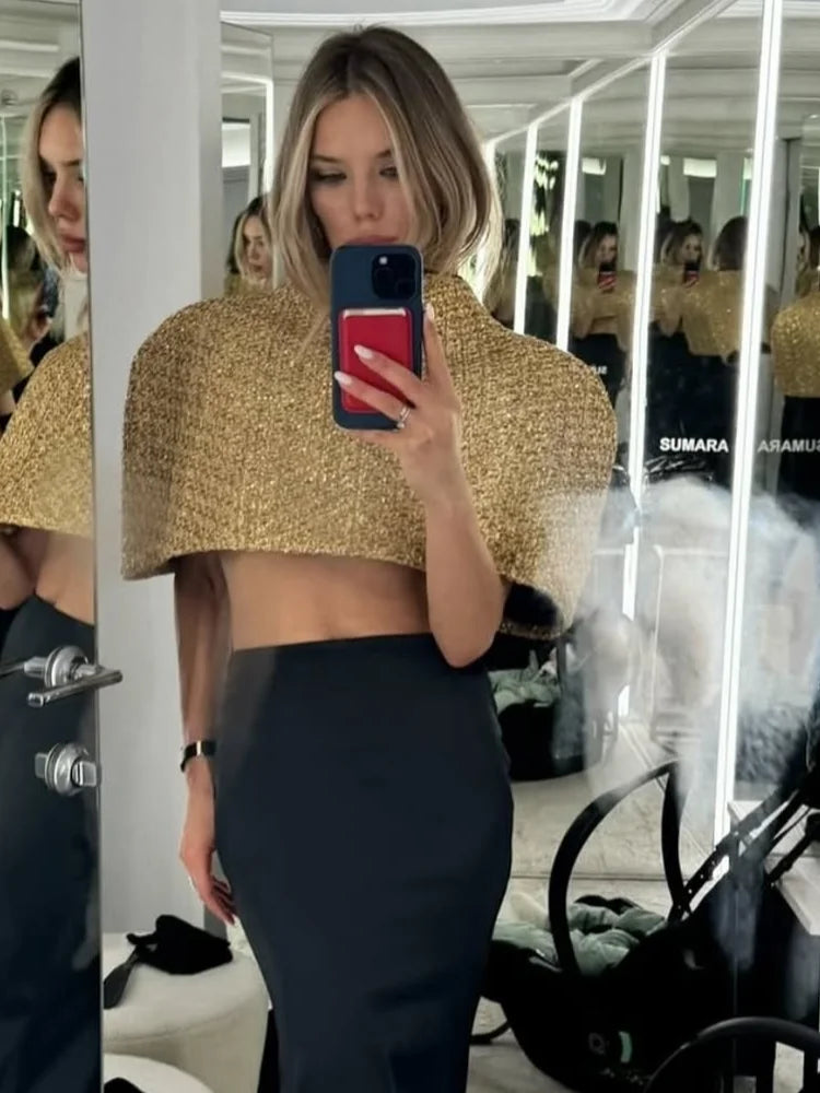Mirror selfie wearing a gold sparkle cape-sleeve top with a high neck, styled with a fitted dark skirt in an indoor setting.