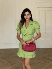 Woman in a Green pleated skirt and blazer set styled with a red bag against a white panelled wall.