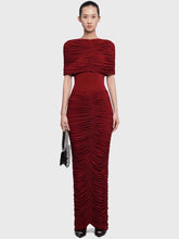 Model wearing a high neck ruched maxi dress with a cape overlay, standing front-facing with a small handbag on a studio backdrop.