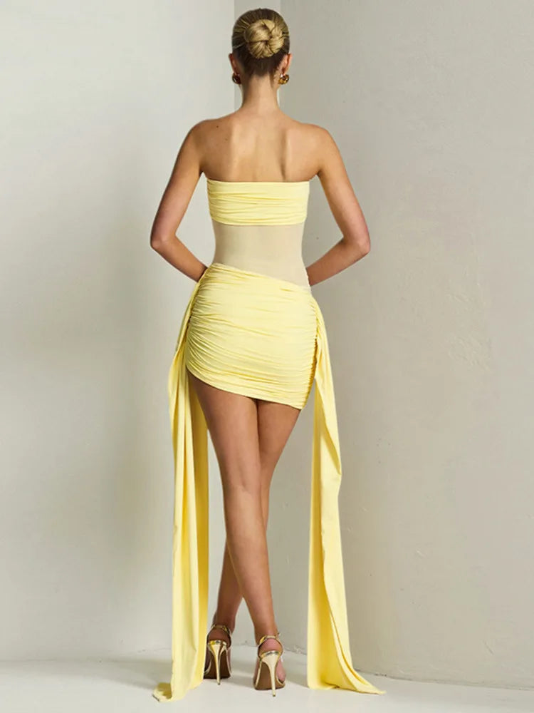 Back view of a woman wearing a yellow one-shoulder dress with a long slit on a plain background
