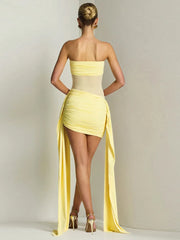 Back view of a woman wearing a yellow one-shoulder dress with a long slit on a plain background