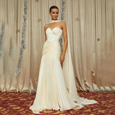 Model in an ivory strapless pleated maxi gown, standing before a curtain backdrop, with a softly draped waist and flowing hem.