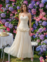 Woman in an ivory strapless long dress standing before a colorful flower wall, showing a draped bodice and airy A-line skirt.