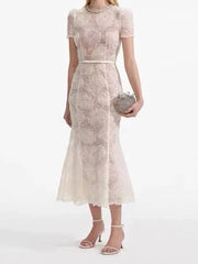 A woman modeling a lace mini dress with a belted waist and a round neckline.