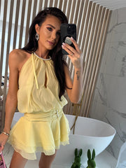 A woman is taking a mirror selfie wearing a light yellow halter mini dress in a modern bathroom.