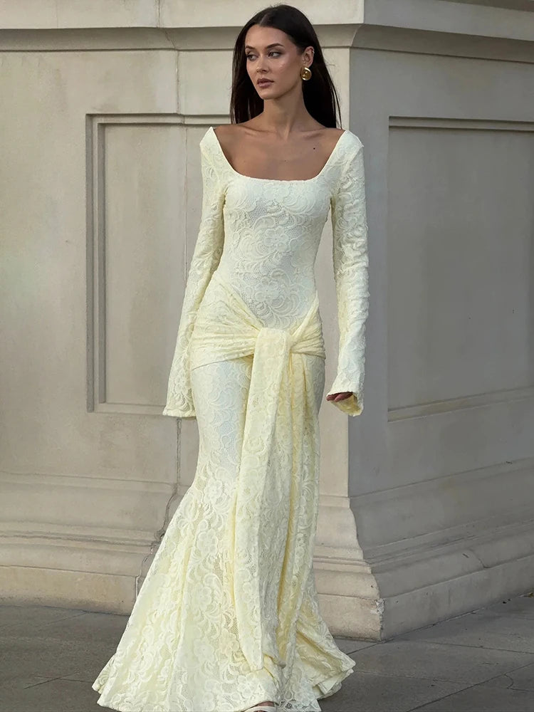 Model wearing a floor-length long sleeve maxi dress with a draped silhouette, standing against a neutral architectural backdrop.