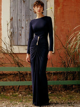 Woman wearing a navy long-sleeve ruched maxi dress standing by a rustic wall