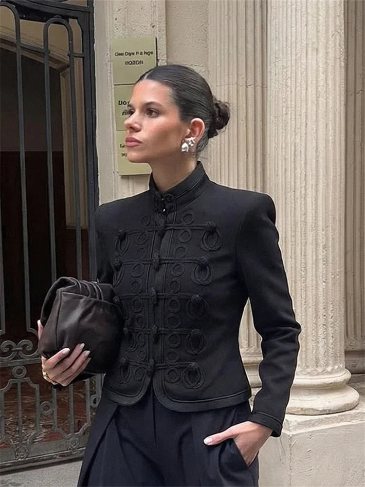 Woman wearing a black mandarin collar blazer with tonal textured detailing, styled with wide-leg trousers in front of a stone column entrance.