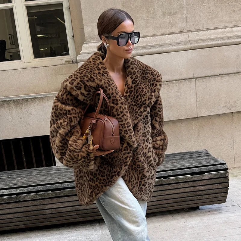 Woman wearing a leopard print coat and sunglasses, holding a brown handbag, standing against a building.